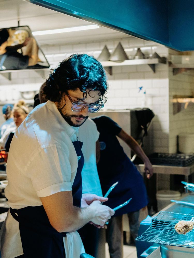 Mohamad Hindi de roupa branca e avental azul cozinhando 