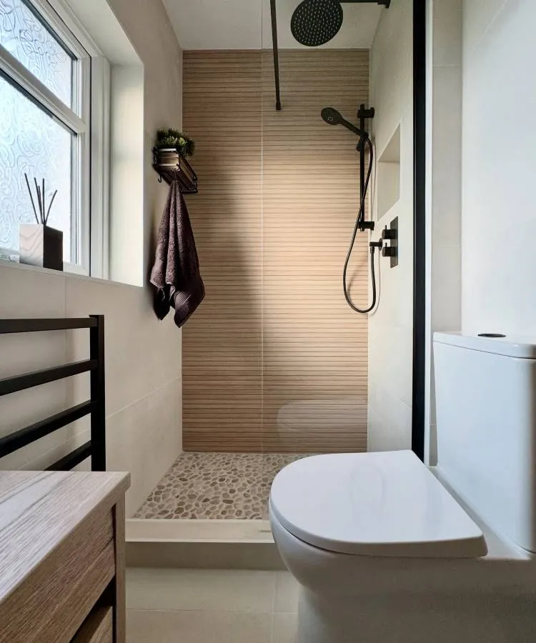 Um banheiro moderno e compacto em tons neutros. Possui um vaso sanitário branco, um chuveiro com azulejos de efeito madeira e piso de seixos, e uma janela com vidro texturizado. Um aquecedor de toalhas preto e uma bancada de madeira clara completam o ambiente.

