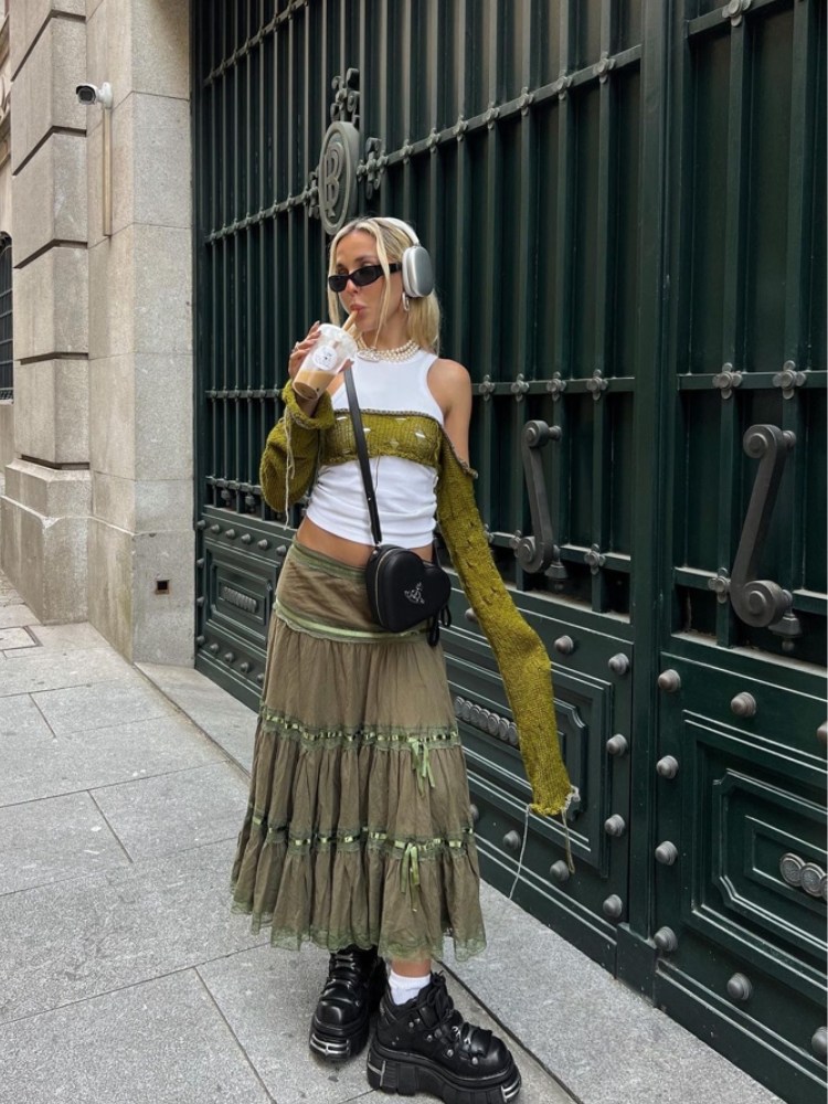 mulher usando regata branca com sobreposição de trico cropped manga longa verde com saia longa em camada verde musgo e oxford de plataforma preto. ela usa cabelo preso, oculos de sol e headfone.