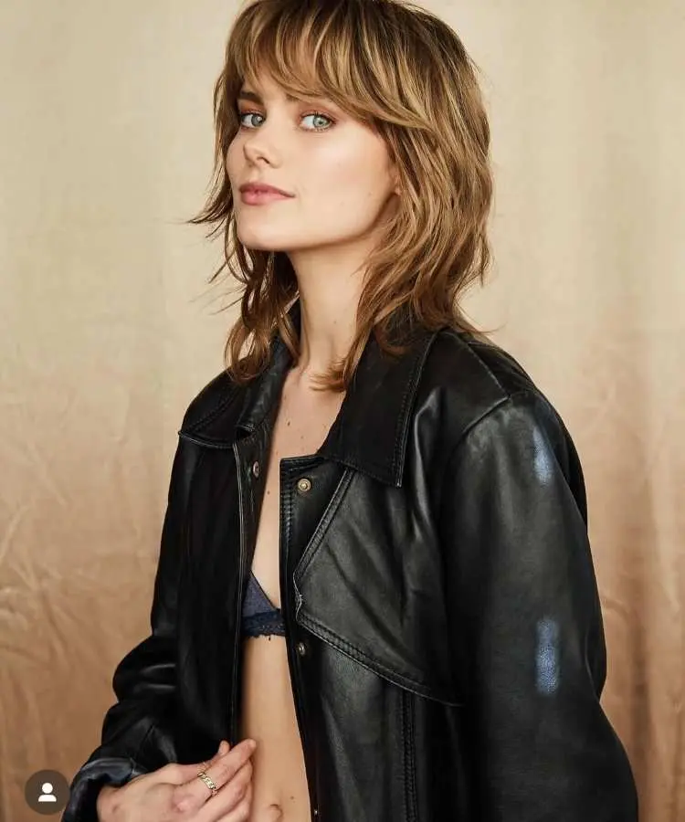 Mulher com cabelo médio loiro acobreado, corte estilo shaggy com camadas desfiadas e franja desconectada. Ela veste jaqueta de couro preta sobre top azul. O fundo é bege neutro, destacando o penteado moderno e despojado.