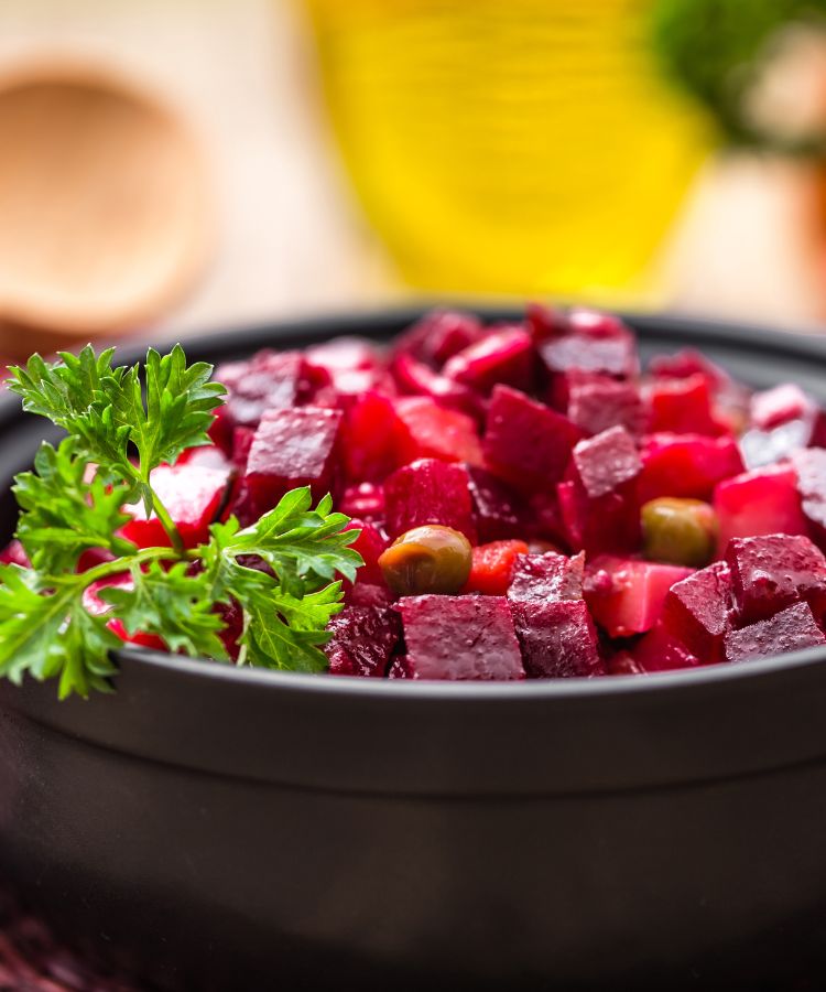 Uma salada de cubos de beterraba e outros vegetais em uma tigela preta. A salada é guarnecida com salsa fresca na borda, com um fundo embaçado e colorido de uma cozinha.