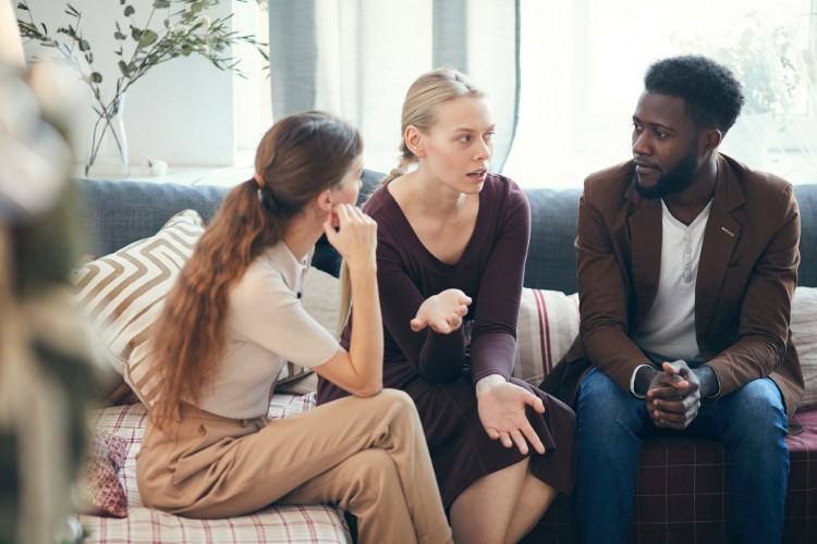 Duas mulheres de pele clara e um homem de pele negra sentados em sofá conversando 