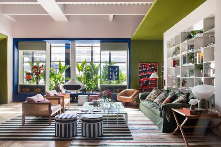 A imagem retrata um interior de sala de estar espaçoso e moderno, com teto verde, paredes brancas e detalhes em azul. Inclui sofás confortáveis, estantes cheias e decoração colorida. Grandes janelas permitem a entrada de luz natural.

