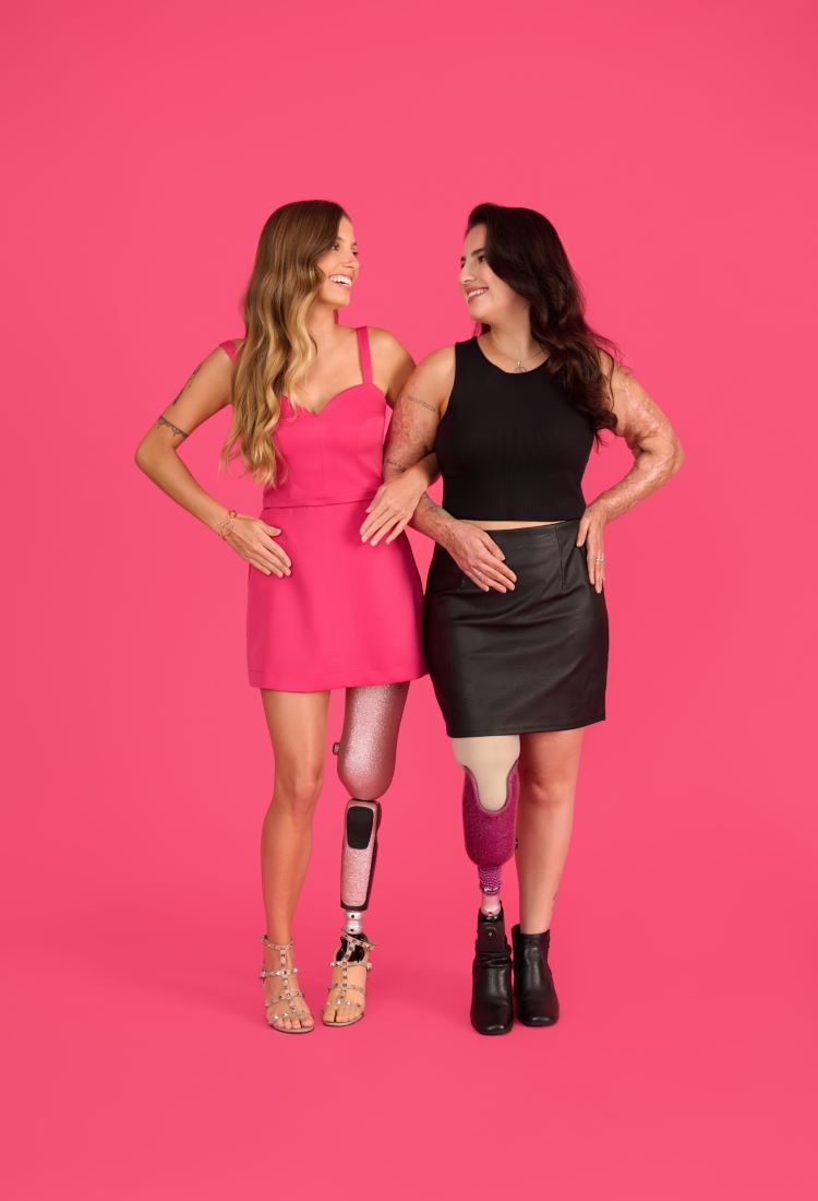 Duas mulheres sorridentes representam as Barbie brasileiras PCDs em frente a um fundo rosa vibrante. Usando próteses transtibiais brilhantes, uma veste vestido rosa com sandálias, outra cropped preto com saia de couro e botas, simbolizando diversidade, inclusão e empoderamento.