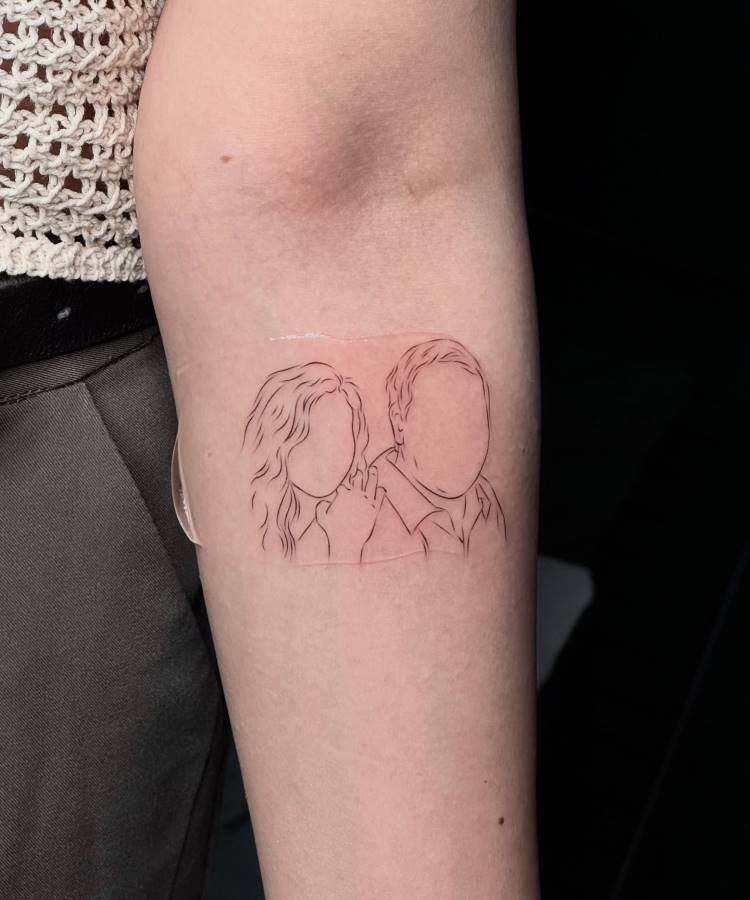 Uma tatuagem de contorno fino no braço exibe as silhuetas de duas pessoas sem traços faciais, uma com cabelo encaracolado e outra com cabelo curto. O desenho minimalista sugere uma conexão pessoal entre os indivíduos.