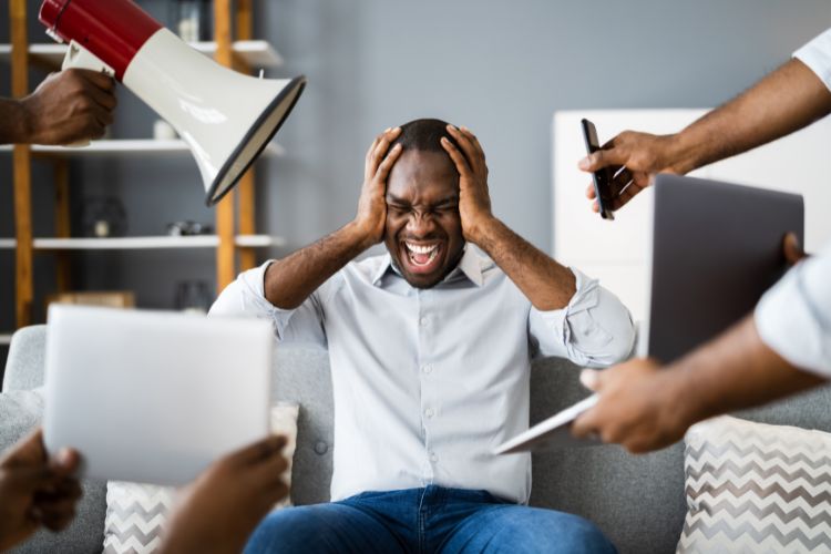 Homem negro incrédulo com as mãos na cabeça perante notícias em dispositivos móveis e auto-falante, segurados por mãos de outras pessoas ao seu redor