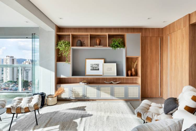 Sala de estar em cinza e madeira, com muita iluminação natural, poltronas off-white e diversos itens decorando a estante, como plantas, vasos e quadros.