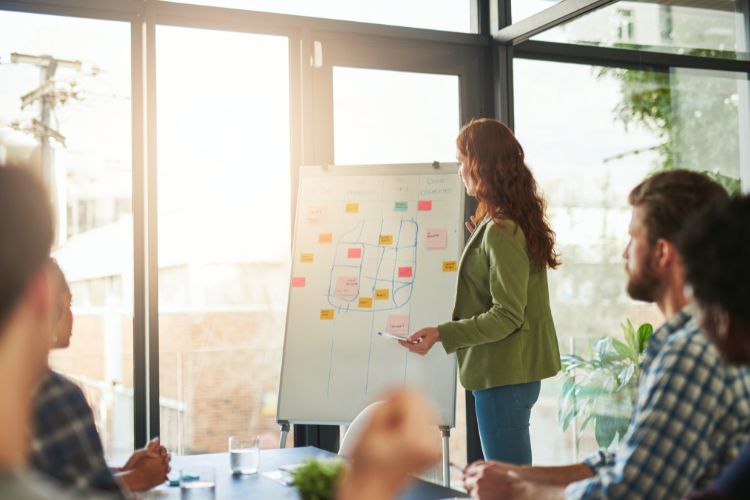 Profissional apresentando ideias em um quadro branco com anotações coloridas, durante reunião em ambiente corporativo iluminado. Discussão sobre estratégias criativas e tendências de publicidade para 2025, envolvendo equipe em colaboração e planejamento para futuros avanços no mercado.
