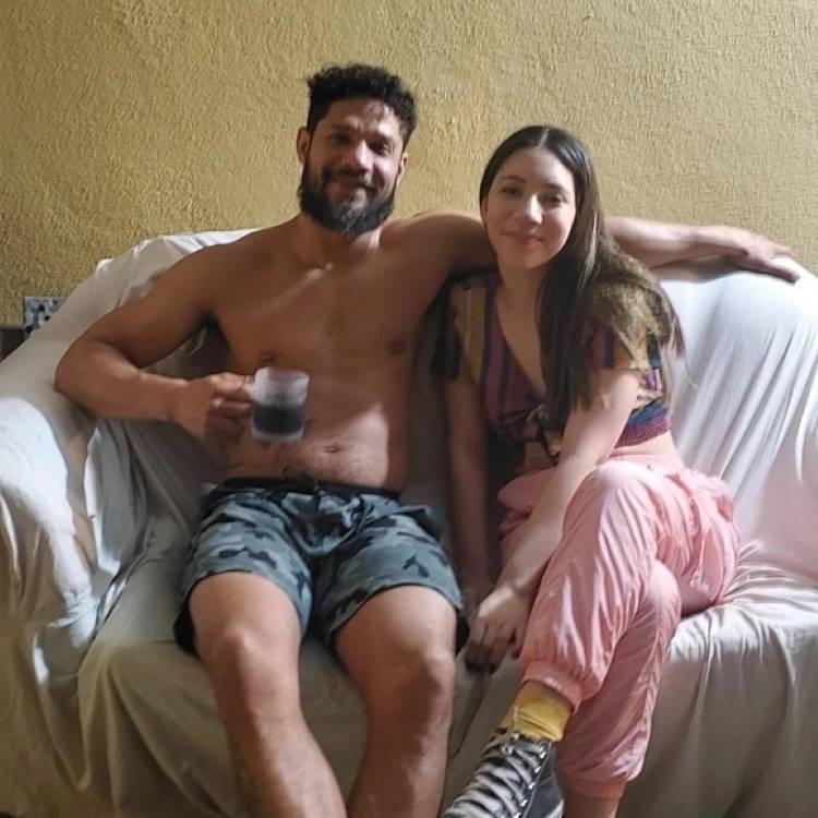Juninho sentado no sofá com capa branca, abraçando sua filha, um jovem de pele clara e cabelo castanho.