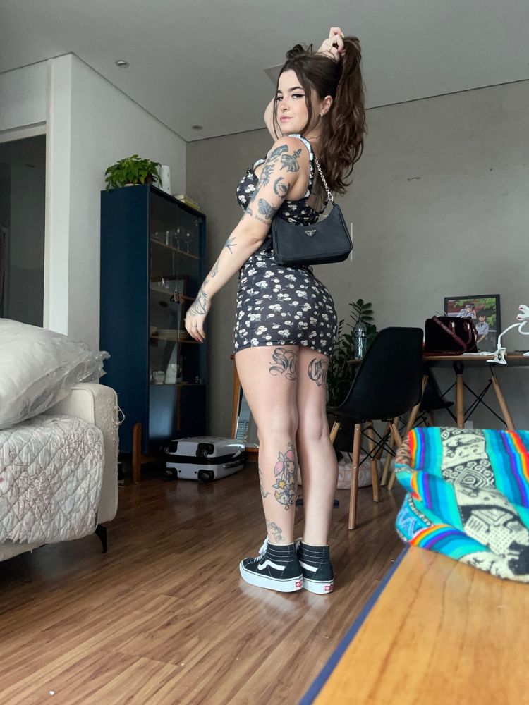 Mulher de pele clara com braços e pernas tatuados, usando vestido curto preto