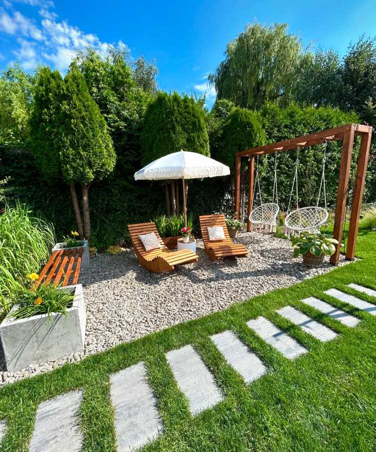 Jardim com chão de brita cercado por grama verde. Duas espreguiçadeiras de madeira com almofadas brancas estão próximas a um guarda-sol. Uma estrutura de madeira suporta balanços de macramê. Plantas e árvores criam um ambiente natural e aconchegante.