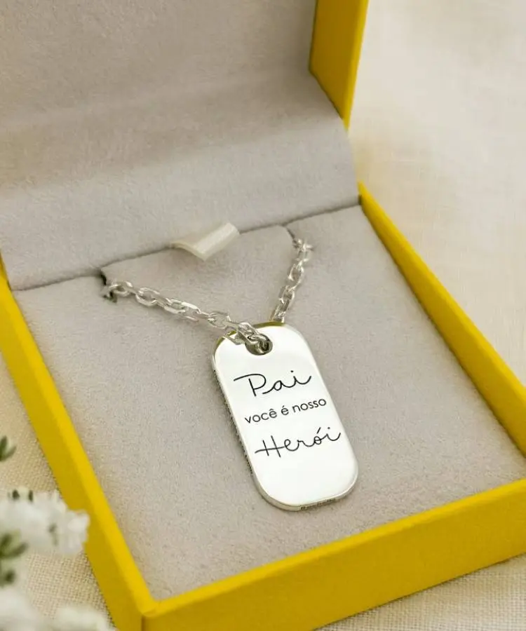 A segunda imagem mostra um colar masculino com uma placa de identificação prateada, gravada com a frase "Pai você é nosso Herói". O colar está delicadamente disposto sobre um forro cinza dentro de uma caixa de presente amarela aberta.







