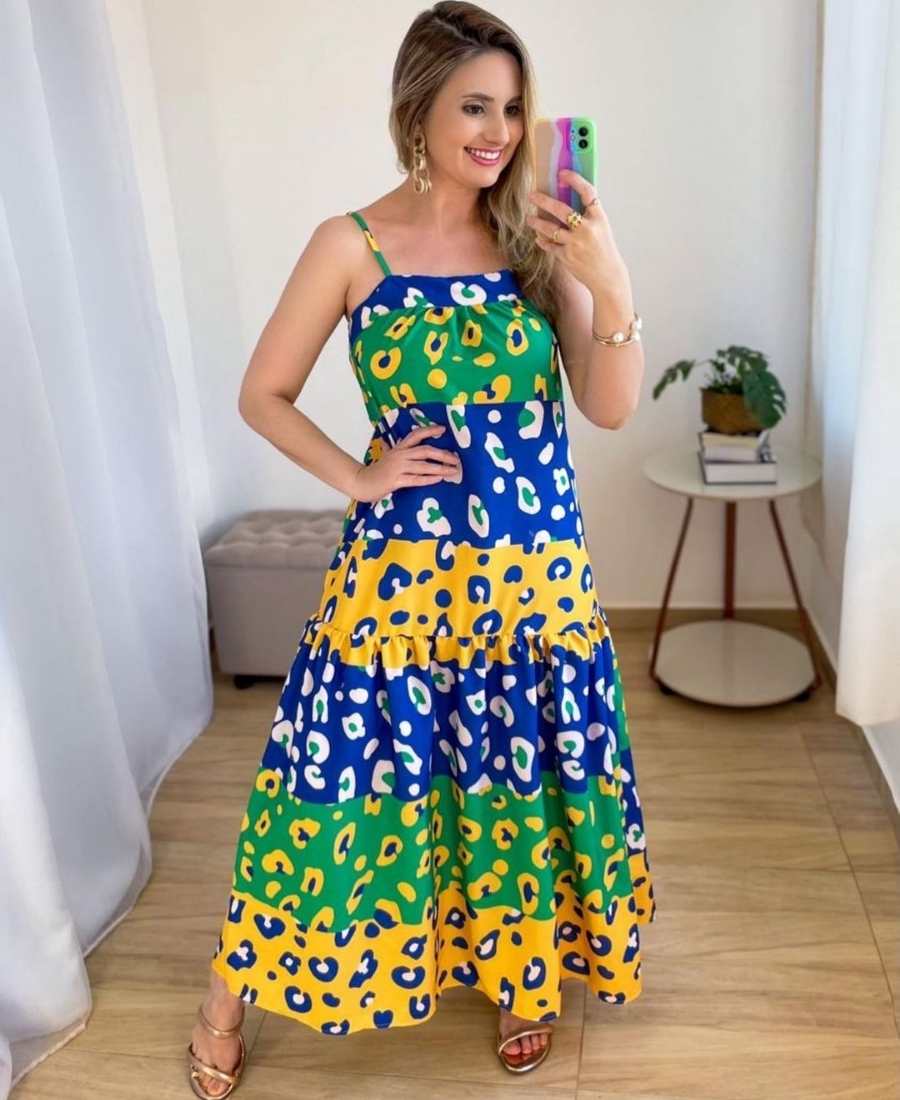 Imagem com fundo de parede, plantas e estofado. No centro, mulher usando roupa copa do mundo: vestido longo com estampa de oncinha.