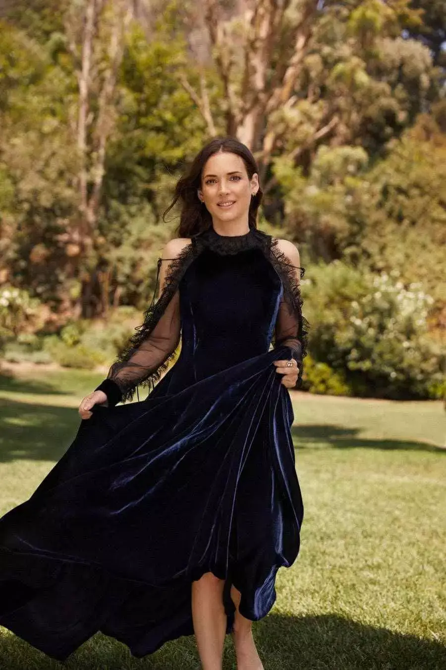 Foto de Wynona Ryder, atriz de Stranger Things, com um vestido longo de camurça, dentro de um parque com muito verde e árvores