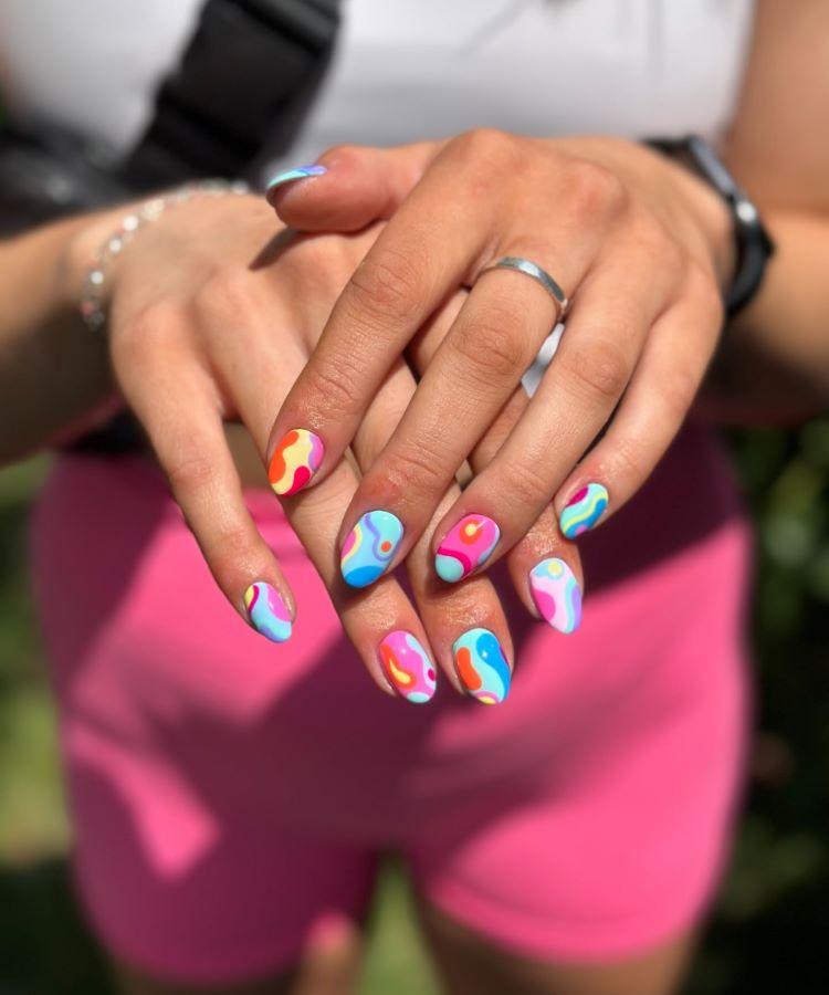 A imagem apresenta unhas de carnaval vibrantes e alegres. Decoradas com formas abstratas em tons de azul, rosa, laranja, amarelo e branco, elas trazem um design psicodélico, criativo e divertido, ideal para celebrar ocasiões festivas e cheias de cor.