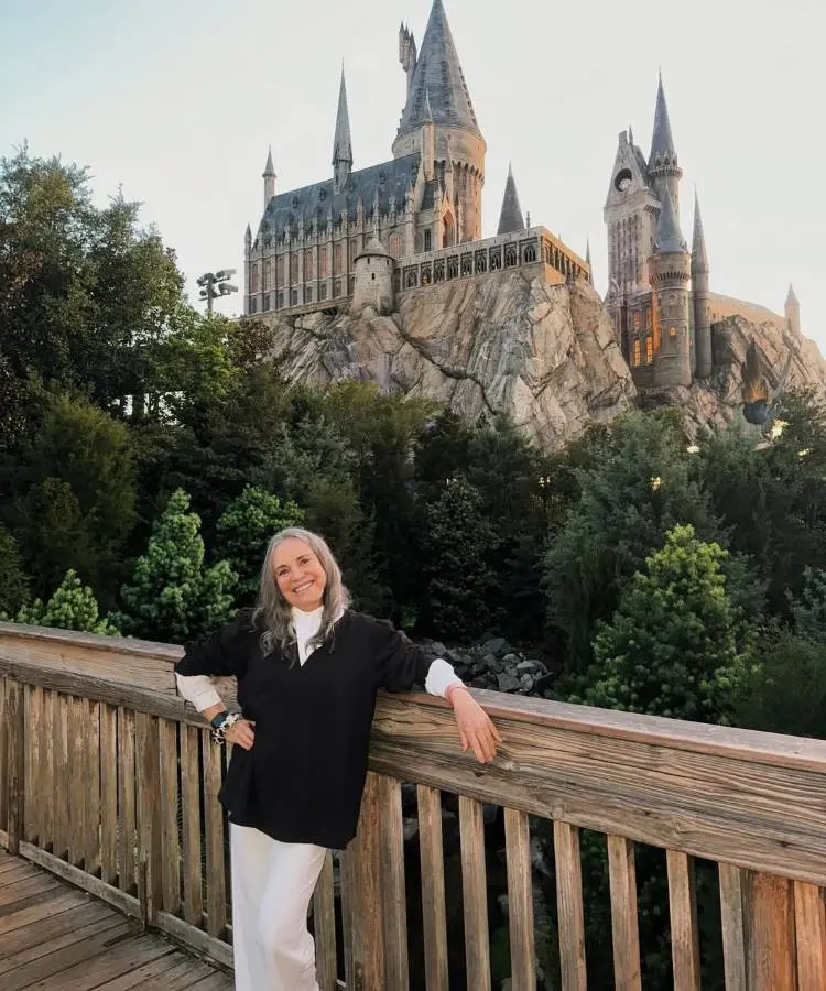 Mulher de cabelos grisalhos longos, usando camisa branca e blusa preta, posa sorridente em frente ao Castelo de Hogwarts, cercado por árvores. Figura marcante do elenco de "Vale Tudo", hoje celebrada por sua personalidade carismática e atuação memorável.