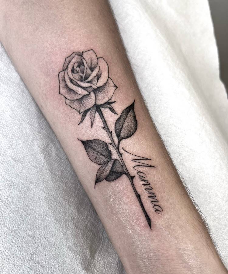 A imagem apresenta uma tatuagem em preto e branco de uma rosa com folhas no antebraço de uma pessoa. O caule da rosa se estende para baixo e tem a palavra "Mamma" escrita em itálico ao longo dele. A tatuagem é detalhada e a pele é clara.







