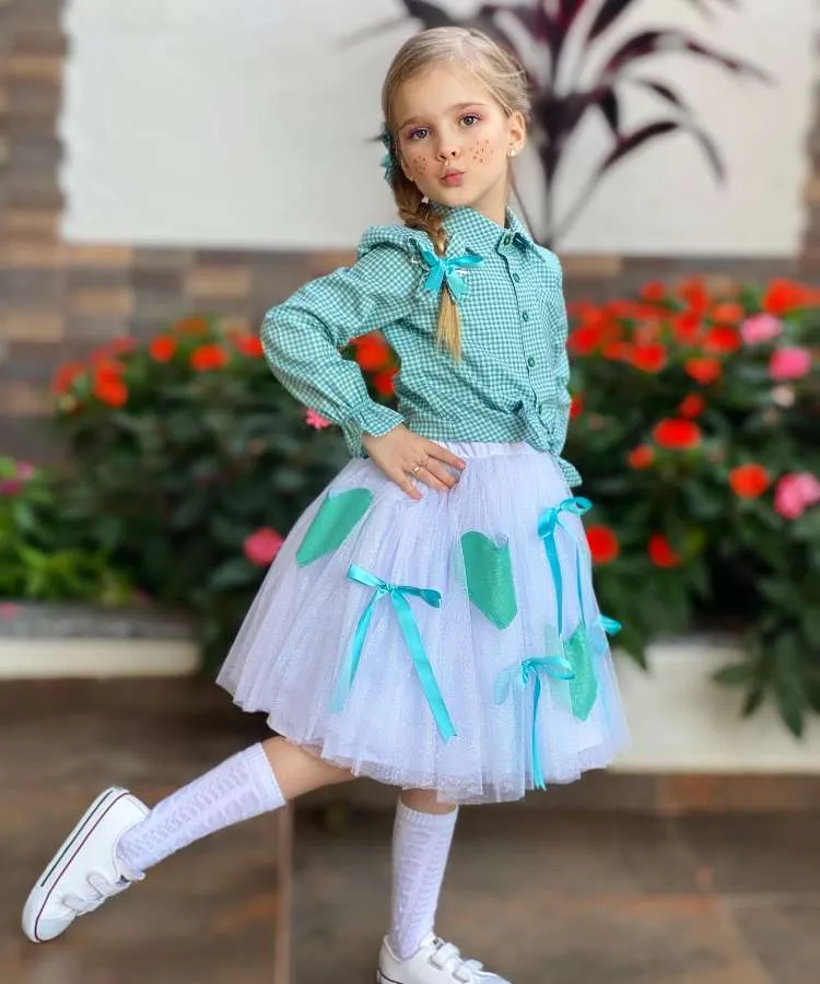Menina usando camisa xadrez verde com saia branca, laços e corações verdes.
