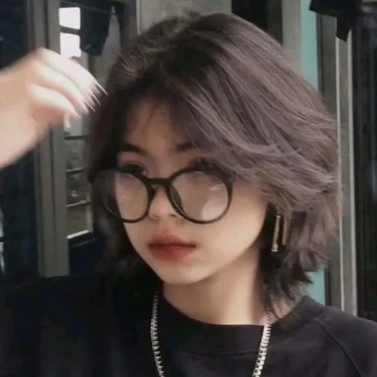 Na foto uma mulher jovem e branca de cabelo preto e curto em camadas. Ela tem o rosto redondo e usa óculos e uma camiseta preta 