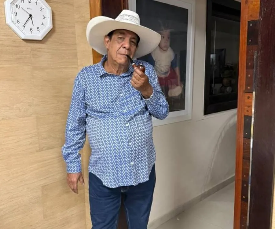Um homem idoso de pele média usa chapéu branco e camisa azul com padrões chevron. Apoia-se contra uma parede beige com relógio e foto emoldurada de criança. Ele segura um cachimbo e o ambiente transmite aconchego e tranquilidade.





