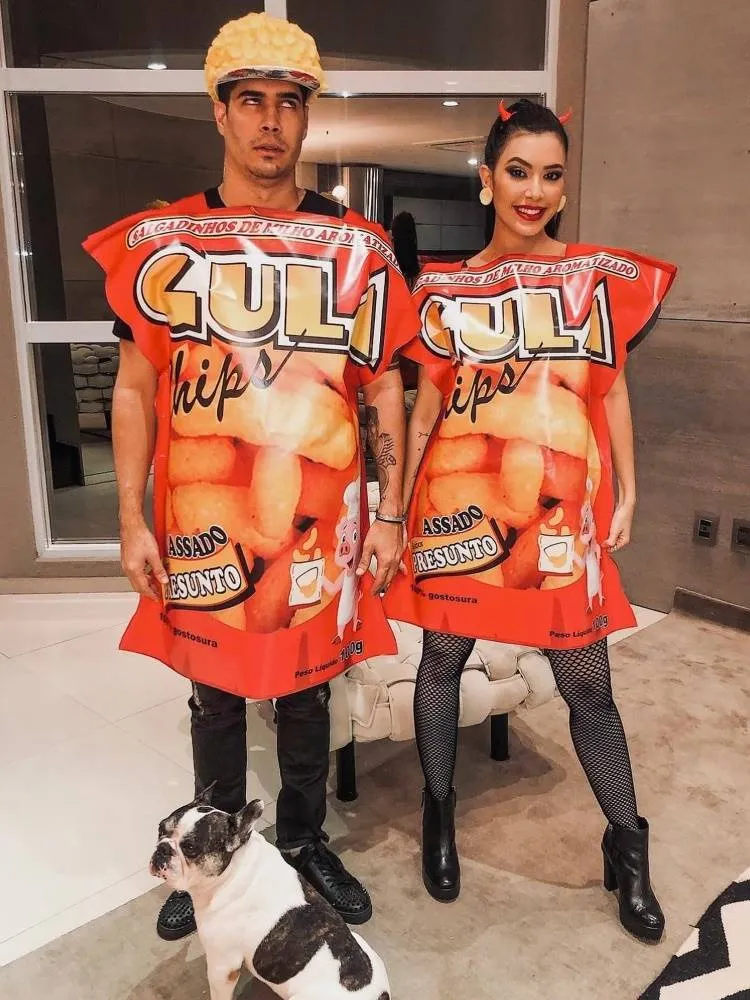 Casal de influenciadores Rica de Marré e Adrão Meron fantasiados de sete pecados capitais (gula) para o Halloween. Ambos usam roupa preta e saco de salgadinho escrito "gula chips"