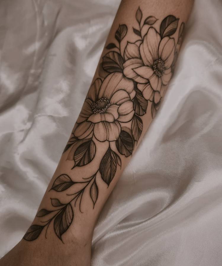 Tatuagem no antebraço com flores grandes e folhas detalhadas em tons de preto e cinza, estilo delicado e sombreado. O braço repousa sobre tecido acetinado branco, destacando o contraste entre a arte da pele e o fundo suave.