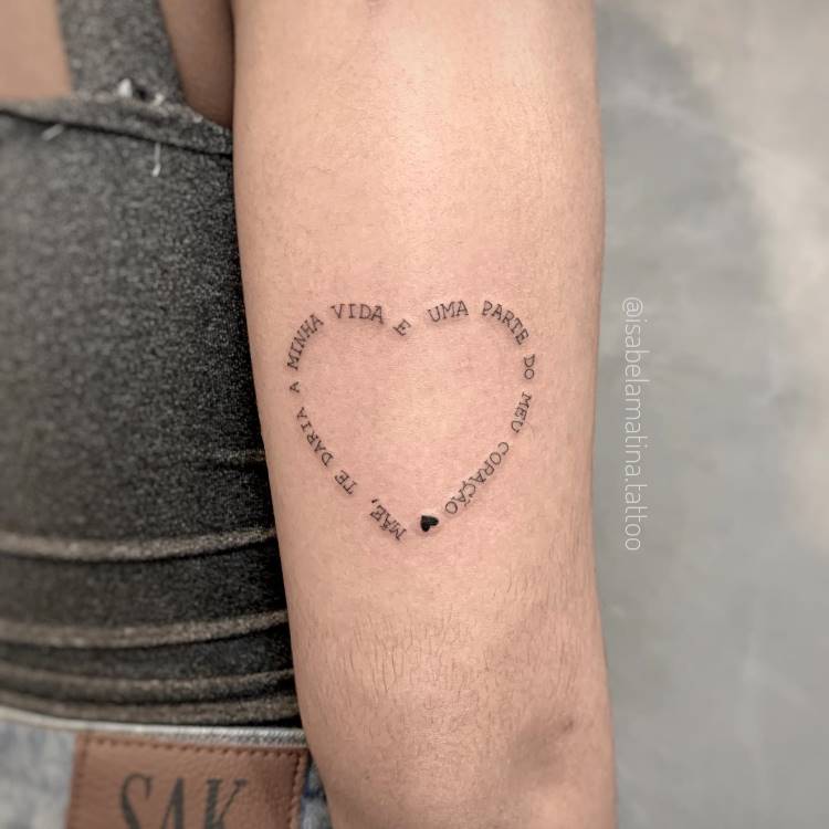 Uma tatuagem de linha fina em formato de coração no antebraço de uma pessoa. As palavras "MÃE, TE DARIA A MINHA VIDA, É UMA PARTE DO MEU CORAÇÃO" estão escritas ao redor do contorno do coração, com um pequeno coração preenchido no final.