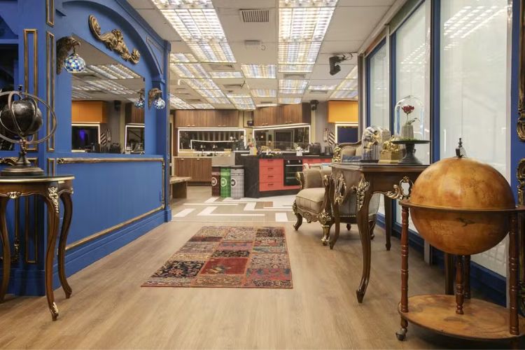 Parte da sala do BBB 24 com parede azul, globo terrestre de enfeite, tapete e vista para as cozinhas. 