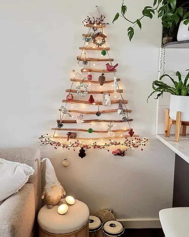 Parede de sala com braço do sofá aparecendo e almofada, no canto, um puff com gnomo, ao lado prateleira com plantas. Na parede, árvore-de-natal de galhos decorados com pelúcias