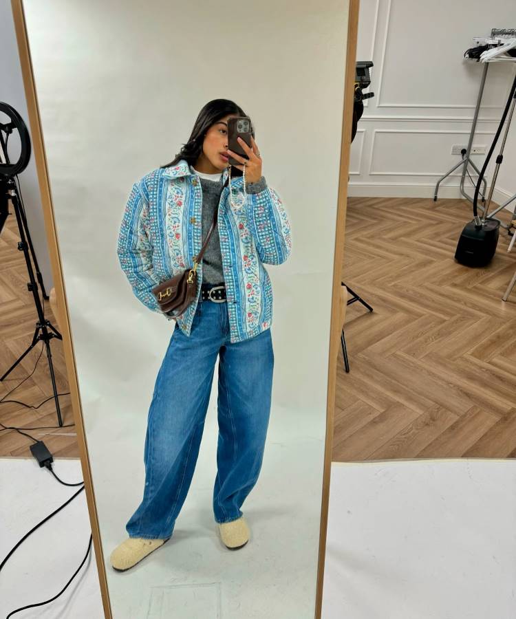 A imagem destaca uma calça jeans barrel leg, tendência de 2025, com cintura alta e corte cilíndrico que proporciona um visual moderno e ousado. A peça combina conforto e estilo, complementando o look casual com jaqueta estampada e acessórios neutros.