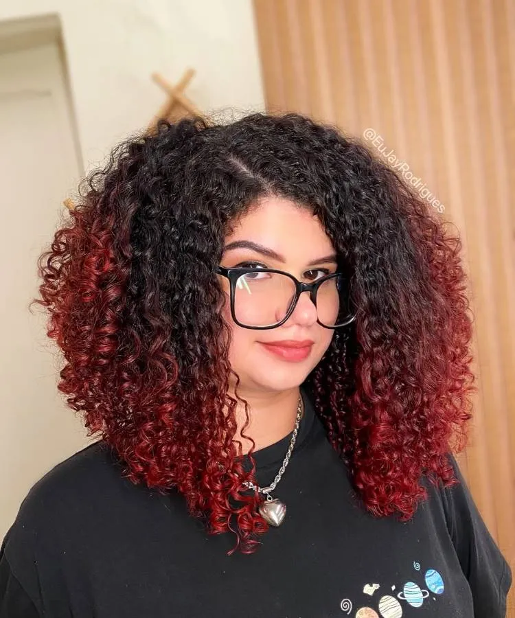 Uma mulher sorri para a câmera com um corte de cabelo cacheado escuro que se degrada para pontas vermelhas vibrantes. Ela usa óculos grandes, um colar de prata e uma camiseta escura.