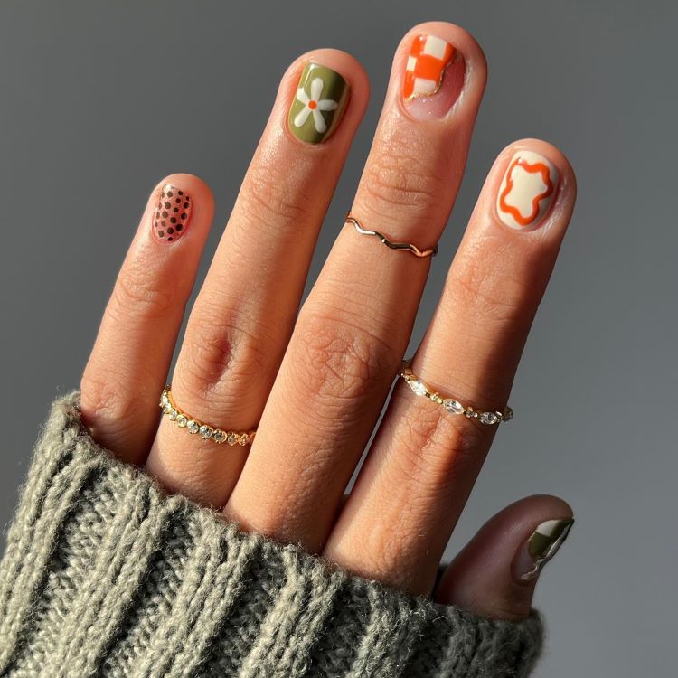 Mão destacando estilos de unhas curtas, cada unha com design único: bolinhas pretas sobre nude, flor branca com fundo verde, xadrez laranja e branco, contorno abstrato laranja. Dedos adornados com anéis dourados delicados e suéter cinza texturizado ao fundo.
