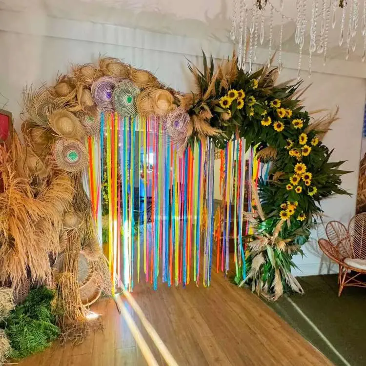 Um arco decorativo para festa Junina, adornado com chapéus de palha, folhagens secas, e girassóis. Fitas coloridas pendem do centro do arco, criando uma entrada festiva.