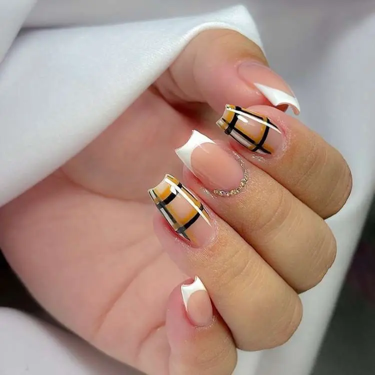 Mão com unhas quadradas e design moderno. Duas unhas têm francesinha branca angular e duas apresentam um padrão xadrez em tons de marrom e preto sobre uma base clara. Há detalhes dourados na cutícula.