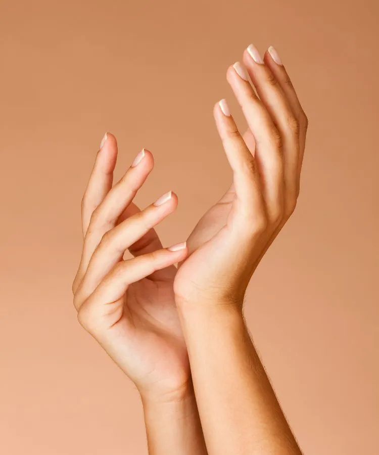 Mãos de mulher com unhas quadradas, com pontas arredondadas, com esmaltação clara. Suas unhas são curtas. As mãos estão em um fundo marrom.
