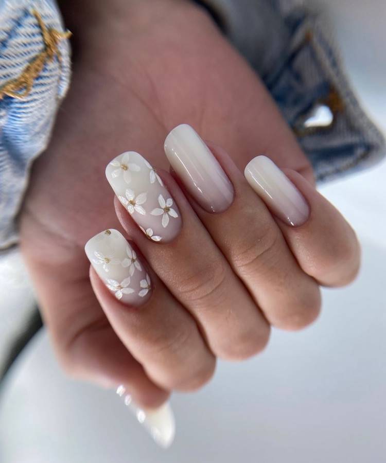 Mão feminina com unhas ovais decoradas com esmalte nude translúcido. Dois dedos apresentam delicados desenhos de flores brancas com detalhes dourados, destacando elegância e sutileza. Fundo desfocado e jaqueta jeans criam um ambiente casual e sofisticado.