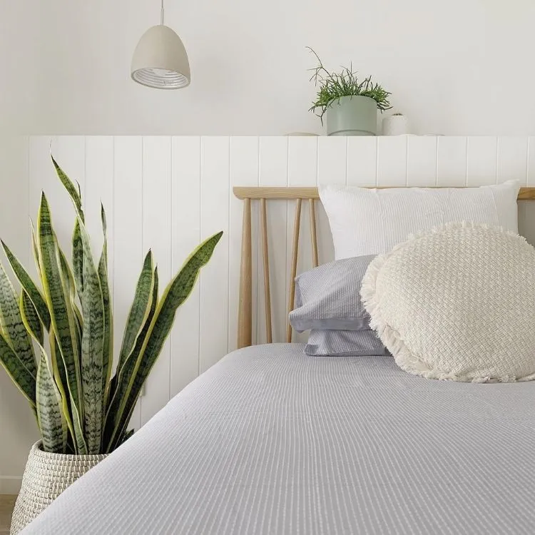 Uma cama com travesseiros e lençóis em azul claro e branco, um vasinho na cabeceira e um vaso grande de lanças-de-Ogum
