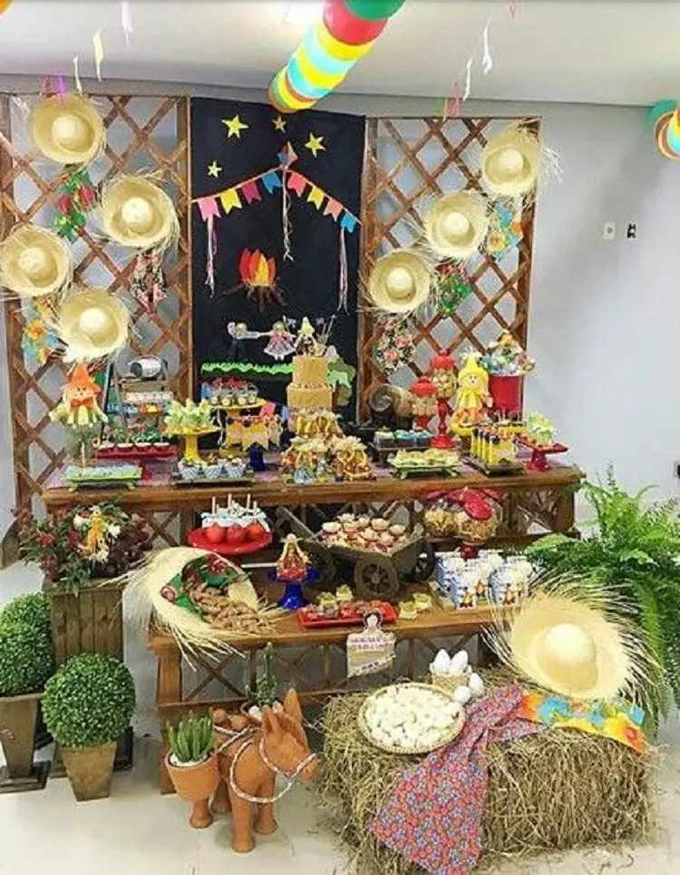 A imagem representa decoração para festa junina. Contém doces e comidas típicas, bandeirinhas, arranjos de flores, chapéus de palha, feno, bexigas, bonecos espantalhos, etc