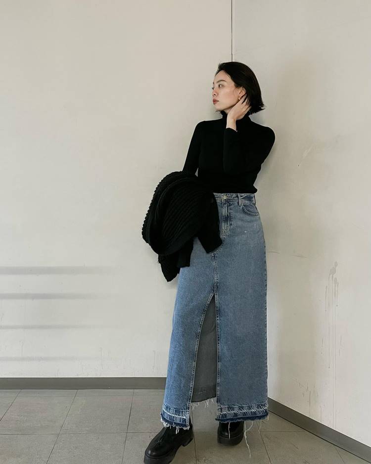 Mulher ocidental usando segunda pele preta, saia longa jeans com fenda no centro, calçado preto, segurando um casaco preto de lã, em uma parede branca.