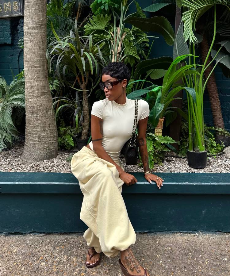Uma mulher com cabelo curto, óculos e tatuagens, sentada casualmente em um muro de jardim. Ela veste uma blusa justa off-white, calças de moletom bege e chinelos marrons. O fundo é composto por plantas tropicais.