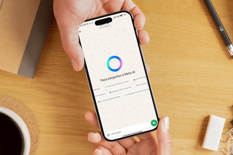 A imagem mostra uma mão segurando um smartphone com a tela aberta no WhatsApp. No centro, há um círculo colorido em tons de azul e rosa, representando a IA do Meta. Abaixo, lê-se "Faça perguntas à Meta AI". O fundo é uma mesa de madeira clara com um caderno, uma caneta e uma xícara de café.



