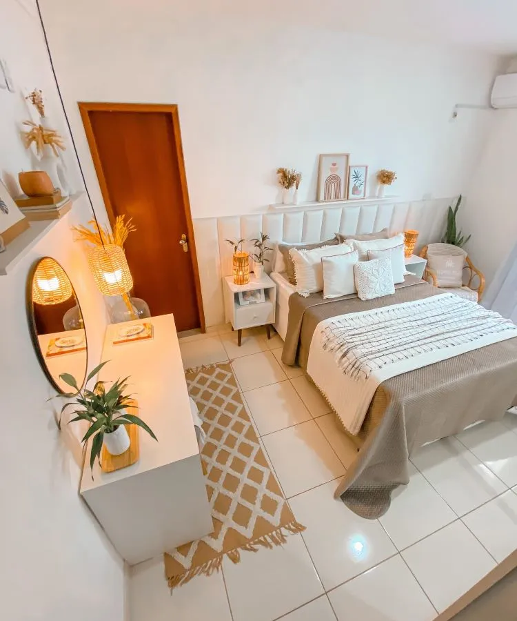 A imagem mostra uma decoração fácil para quarto com cama de casal centralizada, cabeceira cinza com detalhes em arco, almofadas decorativas em tons de cinza e rosa, quadros simples em preto e branco acima da cama, e uma planta verde ao lado sobre um tapete geométrico.

