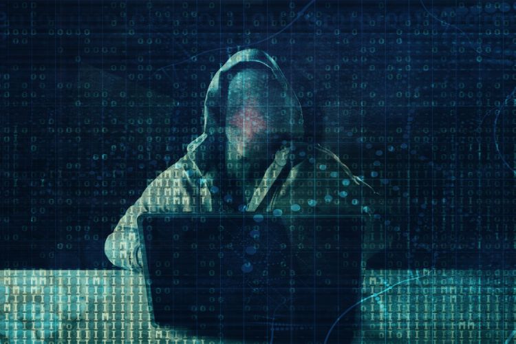 Pessoa sem rosto usando casaco com capuz em frente a um notebook aplicando golpe por inteligência artificial