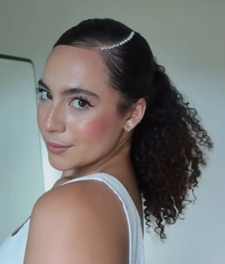 Mulher de pele morena usando cabelo castanho cacheado semi-preso com pérolas