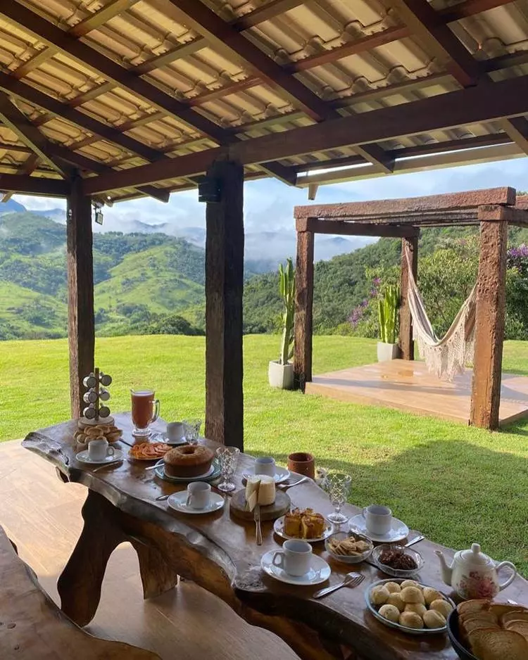 Imagem de fachada de sítio chique e simples. Há uma mesa de madeira esculpida com um café da manhã recheado. Em frente, no jardim de grama, há um pergolado de pedra, com cactos em vasos e uma rede branca no centro