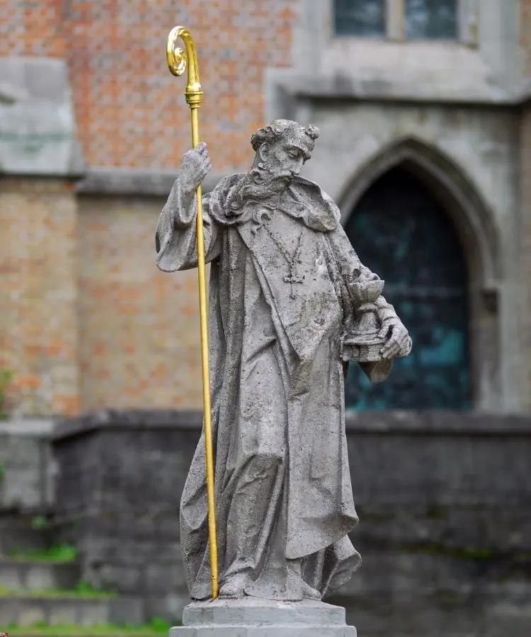Uma estátua de pedra desgastada de um santo (São Bento) barbudo segura um báculo dourado e um objeto em forma de cálice. Ele veste vestes clericais com uma cruz. Ao fundo, uma parede de tijolos e uma porta em arco.