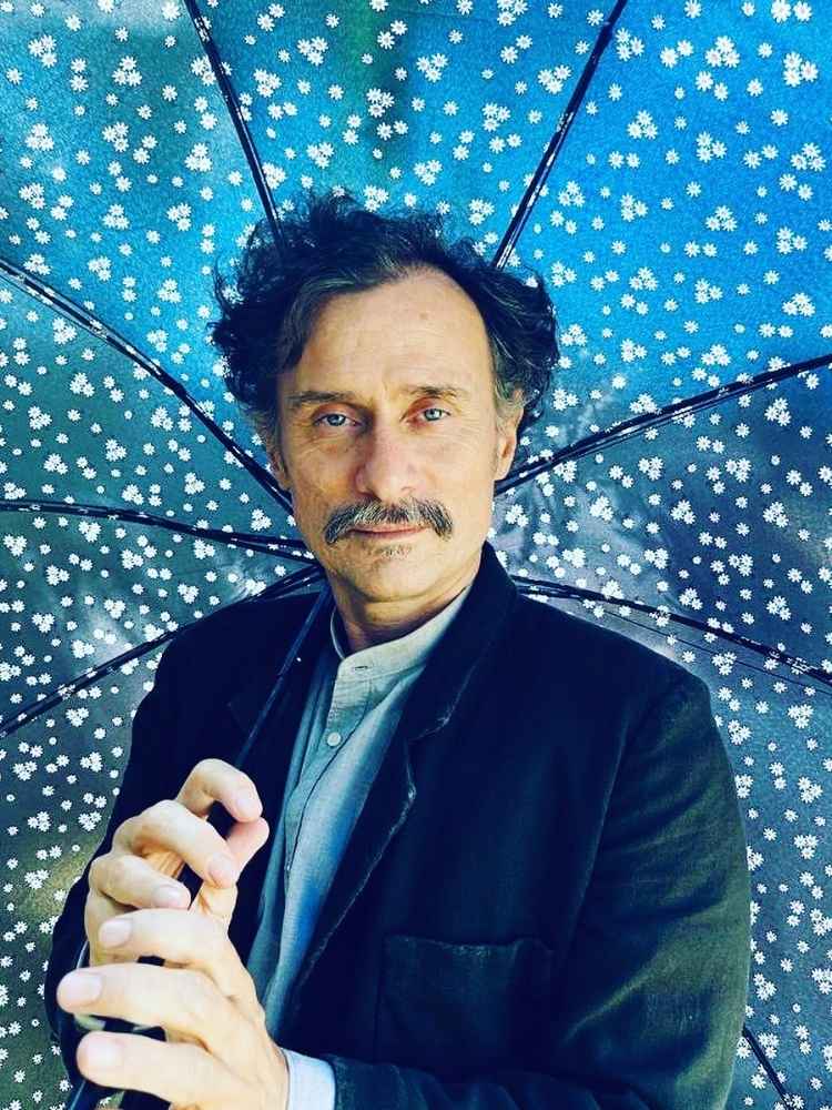 Enrique Diaz com guarda-chuva azul aberto e camisa com blazer preto