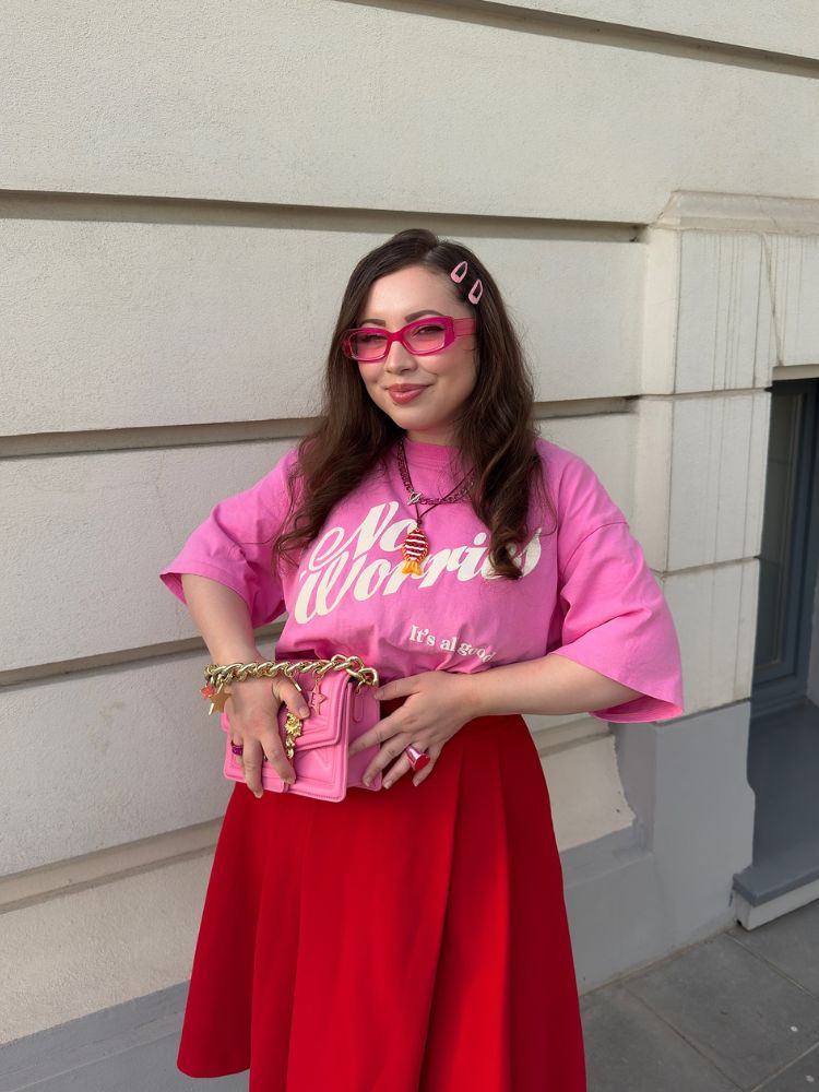 Mulher de pele clara usando óculos de sol com armação rosa transparente e lente rosa, blusa rosa e saia vermelha