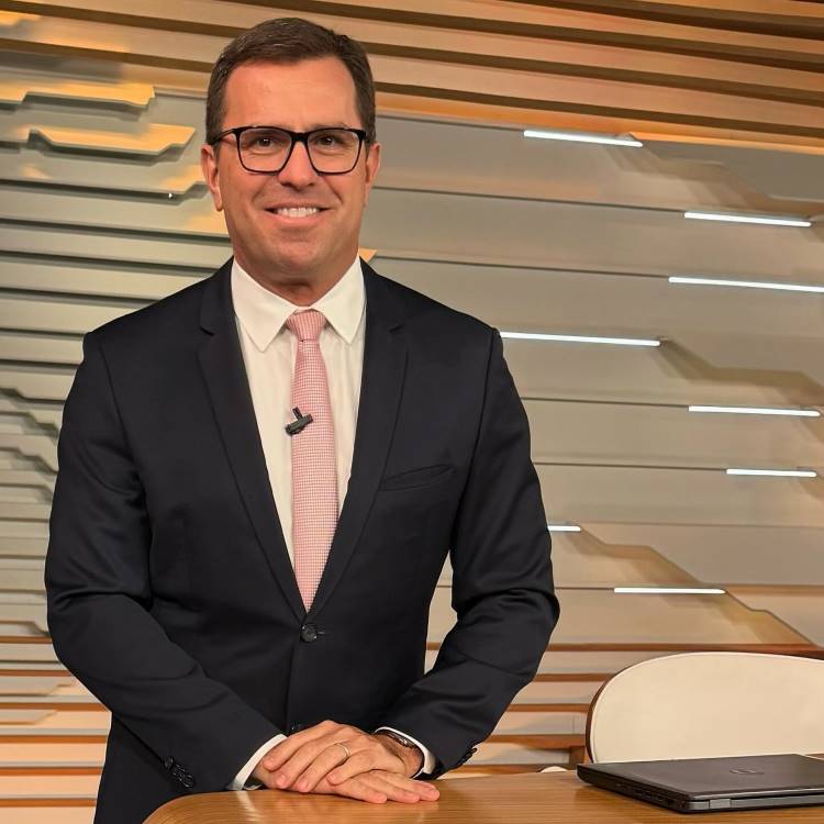 A imagem mostra um homem de terno escuro, gravata rosa clara e camisa branca, sorrindo enquanto está em um estúdio com fundo moderno. Ele está apoiado em uma mesa com um laptop ao lado. para ilustrar o que é compliance.