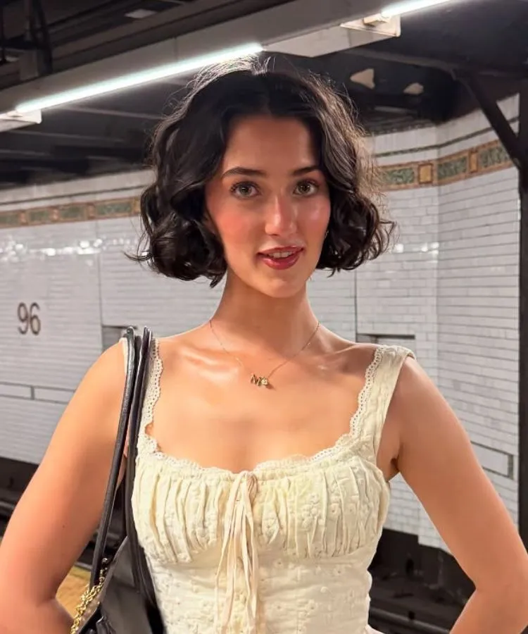 A imagem exibe uma mulher com um corte de cabelo Riviera Bob, um bob castanho escuro e ondulado, em frente a uma parede de azulejos brancos de estação de metrô. Ela sorri levemente e veste um top rendado de cor clara com um pingente em um colar.







